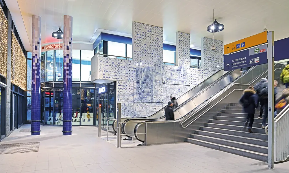 Moderne Bahnhofshalle mit blauen Mosaikfliesen an Wänden und Säulen, einer Rolltreppe, Treppenaufgang und Wegweisern zu U-Bahn und S-Bahn.
