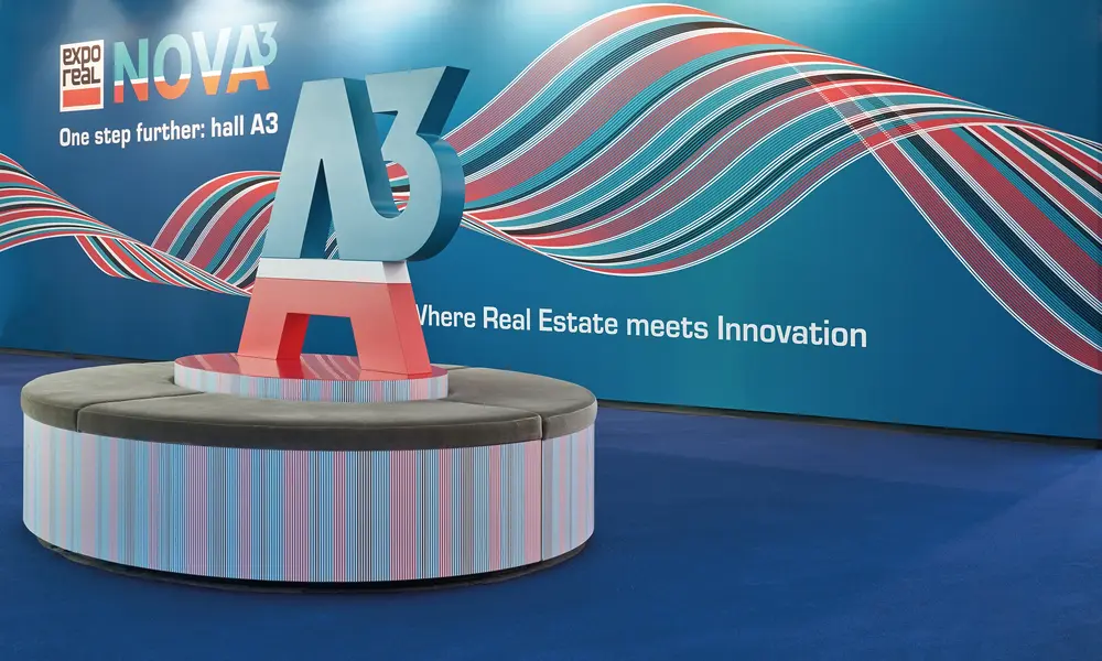 Messestand "expo real NOVA³" mit einer großen "A3"-Skulptur auf einer runden Sitzbank, vor einer blauen Wand mit bunten Wellenmustern und dem Slogan "Where Real Estate meets Innovation".