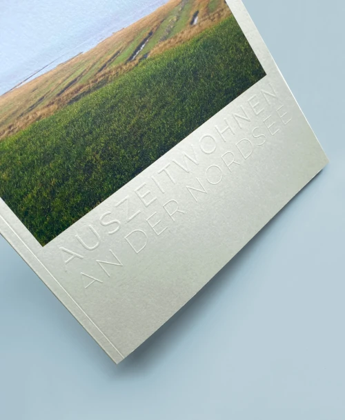 Detailansicht der Broschüre mit eingeprägtem Schriftzug 'Auszeitwohnen an der Nordsee' und einer Landschaftsaufnahme von Wiese und Deich.