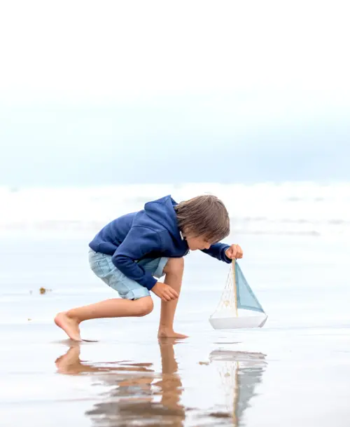 Junge in blauer Kapuzenjacke spielt am Strand mit einem kleinen Segelboot im flachen Wasser.