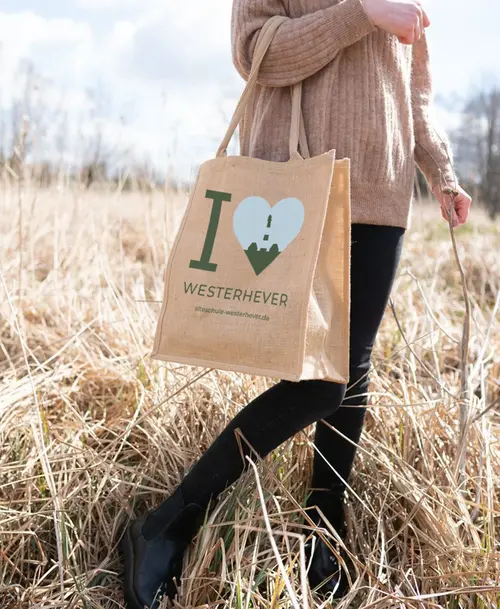 Person mit Jutebeutel mit Aufdruck 'I ❤️ Westerhever' und Logo der Alten Schule Westerhever, stehend auf einer Wiese.