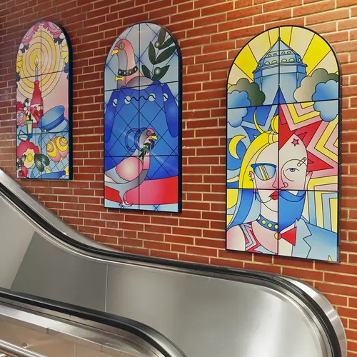 Drei bunt gestaltete, fensterartige Wandbilder mit abstrakten und surrealen Motiven an einer Backsteinwand neben einer Rolltreppe.