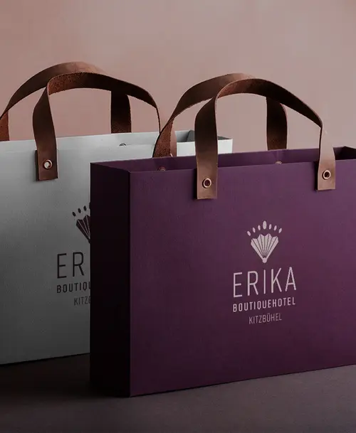 Zwei Einkaufstaschen des Erika Boutiquehotels Kitzbühel, eine in Burgunderrot und eine in Grau, jeweils mit Logo und braunen Ledergriffen.