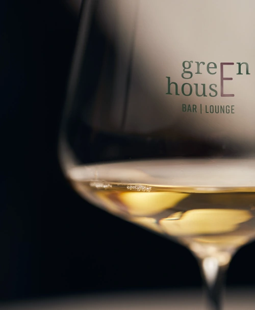 Nahaufnahme eines Weinglases mit goldenem Weißwein, im Hintergrund das Logo der Greenhouse Bar & Lounge.