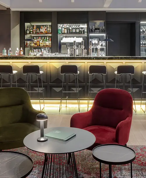 Moderne Bar mit beleuchtetem Tresen, Barhockern und gemütlicher Sitzecke mit roten und grünen Sesseln sowie runden Beistelltischen.