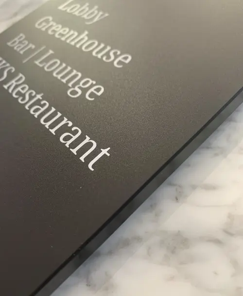 Detailaufnahme eines Schildes im Erika Boutiquehotel Kitzbühel mit weißer Schrift auf schwarzem Hintergrund, das Bereiche wie Lobby, Greenhouse Bar Lounge und Riks Restaurant beschreibt.