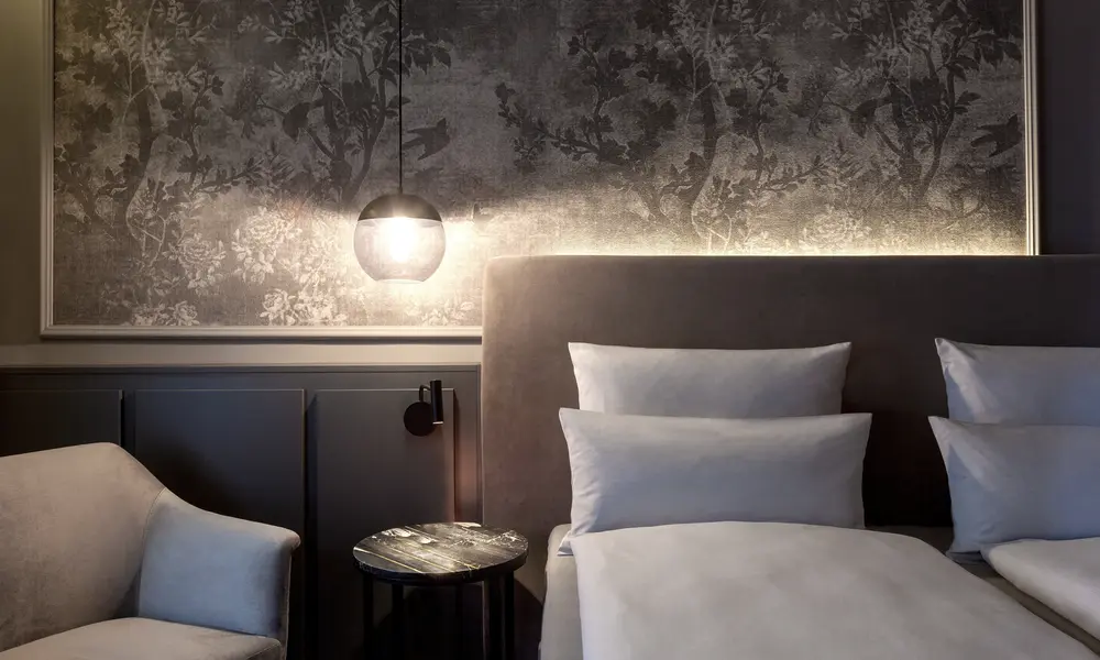 Stilvoll eingerichtetes Hotelzimmer im Erika Boutiquehotel Kitzbühel mit einem Doppelbett, einer gepolsterten Wand mit floralem Muster, einem Sessel und stimmungsvoller Beleuchtung.