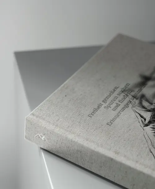 Detail einer edlen Broschüre mit Leinenstruktur, geprägt mit einem Bergmotiv und dem Text 'Freitag genießen, Spuren suchen und bleibende Erinnerungen sammeln'.