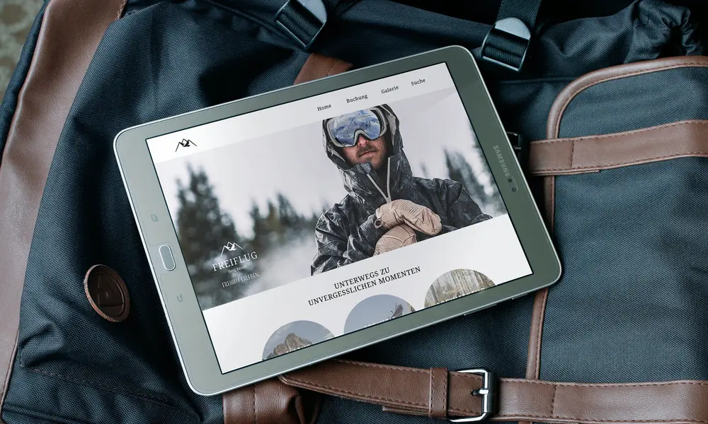 Tablet mit geöffneter Website des 'Freiflug Berg Resort', gezeigt auf einer Reisetasche mit Bild eines Skifahrers und dem Text 'Entdecke die unvergesslichen Momente'.