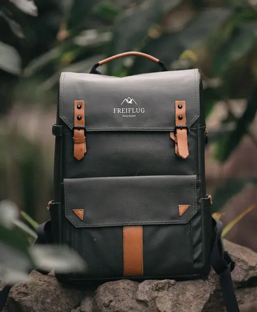 Eleganter dunkelgrüner Rucksack mit braunen Lederriemen und dem Logo des 'Freiflug Berg Resort', präsentiert in einer natürlichen Umgebung.