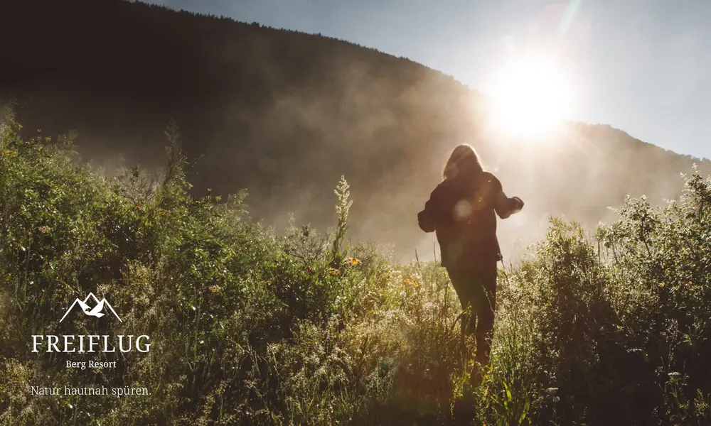Person wandert durch eine sonnenbeschienene Wiese mit Bergen im Hintergrund, begleitet vom Logo des 'Freiflug Berg Resort' und dem Slogan 'Natur hautnah spüren'.