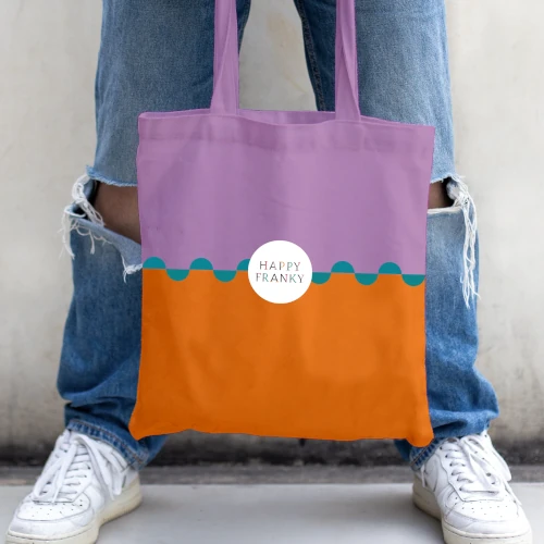 Bunte Stofftasche in Lila und Orange mit wellenförmigem blauen Detail und rundem Logo "HAPPY FRANKY", gehalten von einer Person in zerrissenen Jeans und weißen Sneakern.