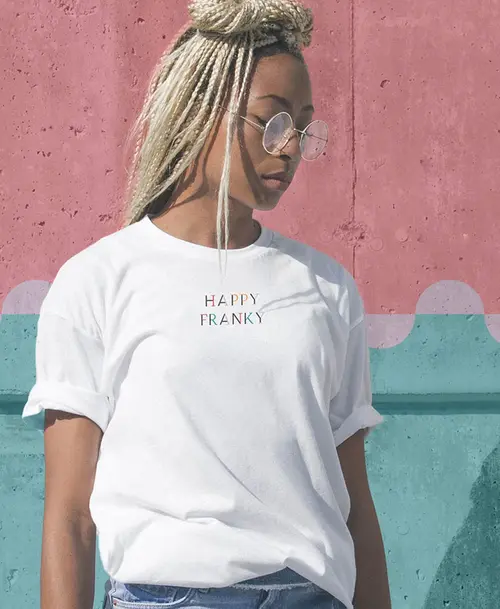 Frau mit weißen T-Shirt, bedruckt mit dem Logo 'Happy Franky', vor einer Wand mit CI-Elementen vom 'Happy Franky'.