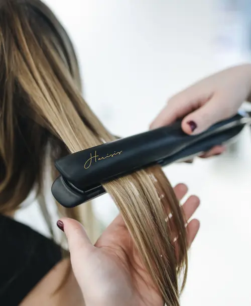 Friseurin glättet blonde Haare mit einem schwarzen Glätteisen, das das Logo von „Harisis – Personal Hairstyling“ trägt.