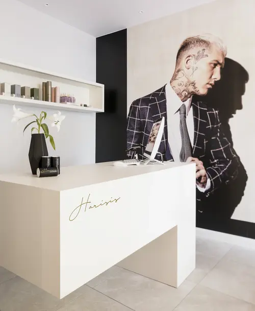 Moderner Empfangsbereich des Friseursalons „Harisis – Personal Hairstyling“ mit cremefarbenem Tresen, Wandregal mit Pflegeprodukten und großem Wandbild eines tätowierten Mannes im Anzug.