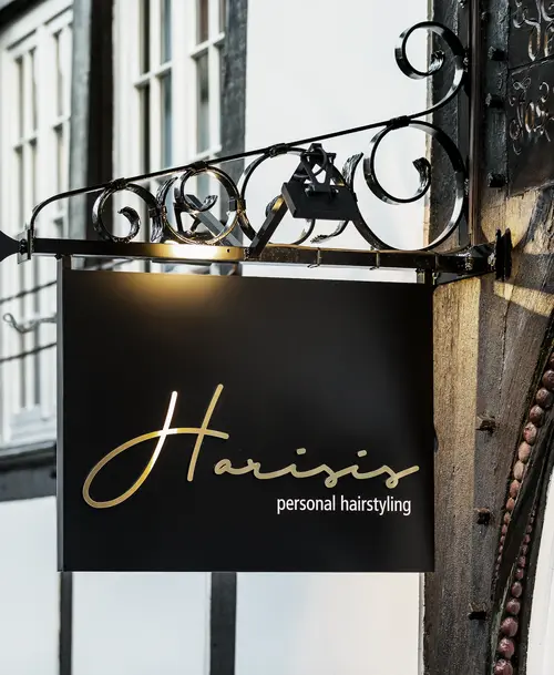 Elegantes schwarzes Firmenschild mit goldener Schrift für „Harisis – Personal Hairstyling“, befestigt an einer verzierten Metallhalterung an einer Fachwerkhausfassade.