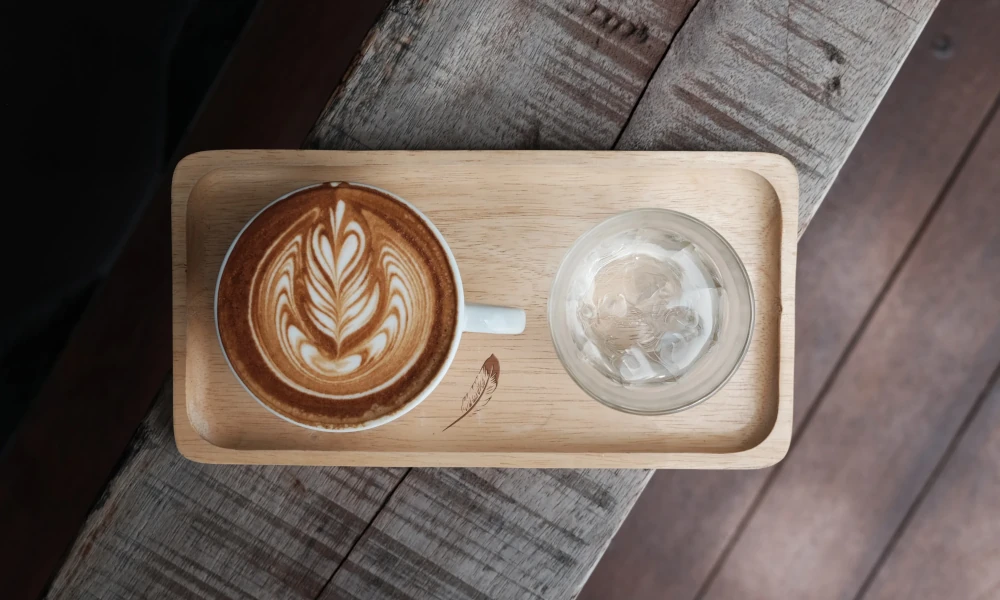 Latte mit kunstvollem Milchschaum und Glas Wasser auf einem Holztablett mit Federlogo – stilvoll serviert auf rustikalem Holztisch im HORST Berg Café.