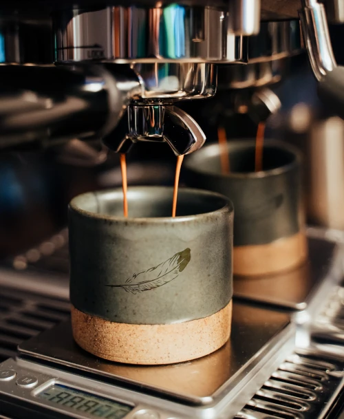 Espressomaschine brüht frisch aufgefangenen Kaffee in rustikale Keramiktassen mit Federlogo – hochwertige Zubereitung im HORST Berg Café mit Fokus auf Handwerk und Qualität.