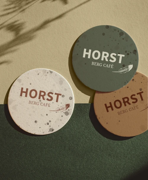 Drei runde Untersetzer mit HORST Berg Café Logo in Naturfarben – stilvolle Markenaccessoires mit Sprenkeldesign und Federmotiv, ideal für ein modernes, umweltbewusstes Café-Ambiente.        ChatGPT fragen