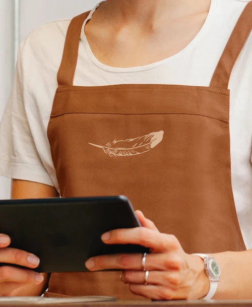 Mitarbeiter:in im HORST Berg Café mit brauner Schürze und Federlogo, hält ein Tablet – modernes, serviceorientiertes Auftreten mit natürlichem Markenstil.