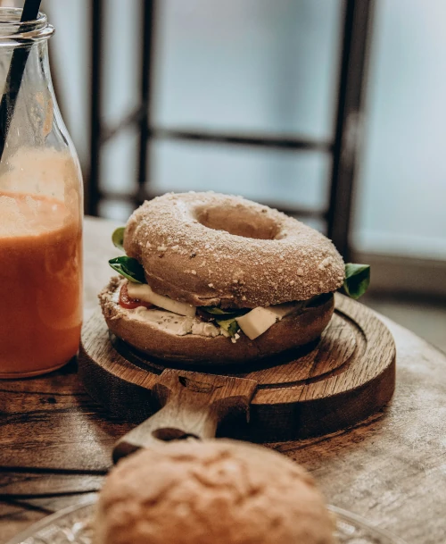 Frisch belegter Vollkorn-Bagel mit Salat und Käse auf Holzbrett, daneben eine Flasche Karottensaft – gesunde, liebevoll präsentierte Frühstücksauswahl im HORST Berg Café.