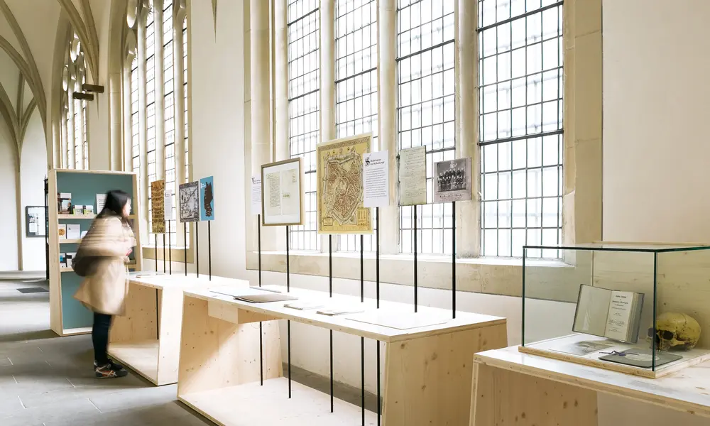 Ausstellungsraum mit hohen Fenstern, Holztischen und Infotafeln, die historische Dokumente und Karten zeigen. Rechts eine Vitrine mit einem Totenschädel. Eine Besucherin bewegt sich durch die Ausstellung.