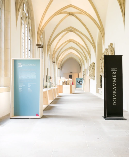 Lichtdurchfluteter Ausstellungsraum mit gotischen Gewölbedecken, Informationstafeln und Holzvitrinen. Im Vordergrund eine blaue Texttafel und eine schwarze Stele mit der Aufschrift „DOMKAMMER“.