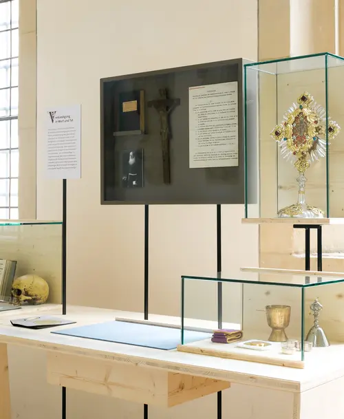 Vitrinen in einer Ausstellung mit religiösen Artefakten, darunter ein vergoldeter Reliquienschrein, ein Kelch, ein Kruzifix und historische Dokumente. Links eine Vitrine mit einem Totenschädel.