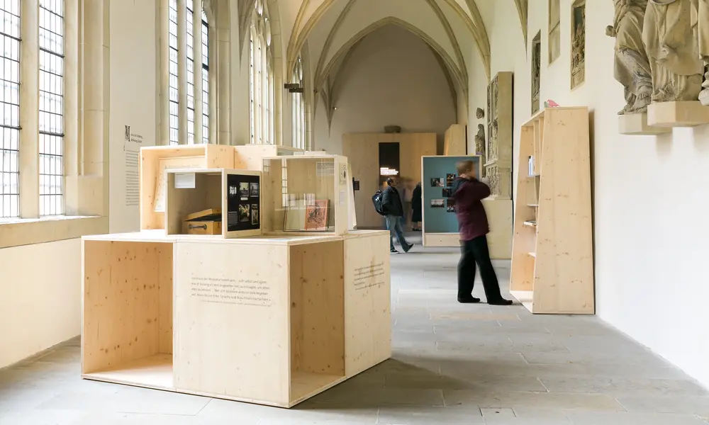 Ausstellungsraum mit gotischen Gewölbedecken und modernen Holzpräsentationen, die historische Dokumente und Artefakte zeigen. Zwei Besucher betrachten die Exponate.