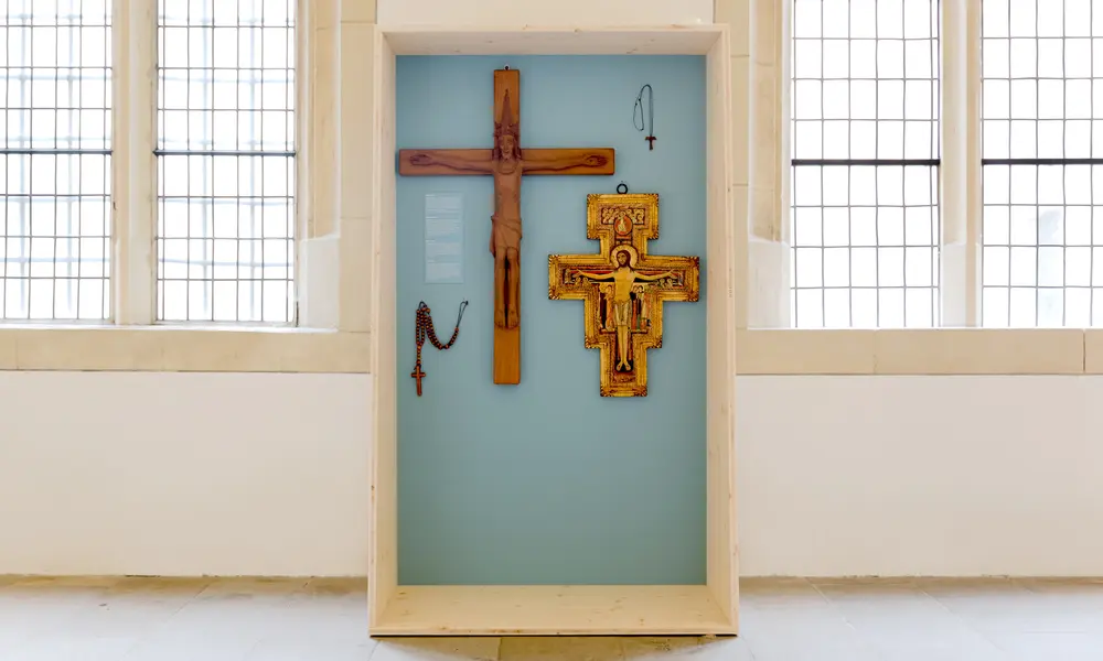 Holzvitrine mit blauem Hintergrund in einer Ausstellung, darin zwei Kreuze, ein Rosenkranz und ein Anhänger, präsentiert vor großen Fenstern.