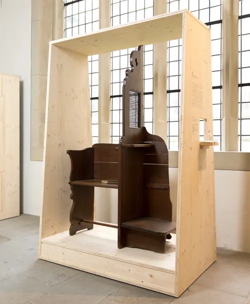 Historischer Beichtstuhl aus dunklem Holz in einer schlichten Holzvitrine, ausgestellt vor großen Fenstern in einem lichtdurchfluteten Raum.