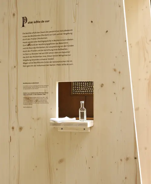 Holzwand mit Informationstafel zur Beichte und einem kleinen Durchlass, durch den eine Flasche Wasser und ein Taschentuch auf einem Regal sichtbar sind.