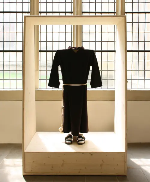 Kapuzinerkutte mit Gürtel und Sandalen in einer Holzvitrine, ausgestellt vor großen Fenstern mit Gittermuster.