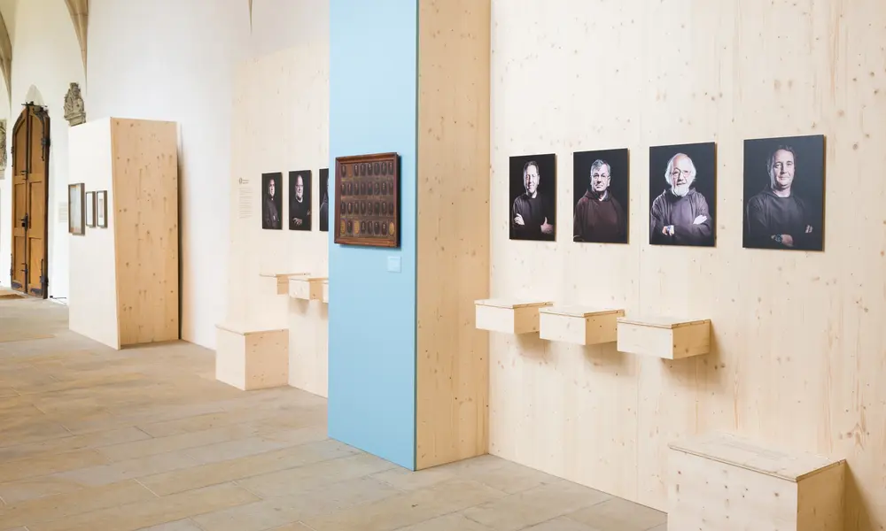 Ausstellungswand mit Porträtfotografien von Kapuzinermönchen, kleinen Holzkisten und einer blauen Informationstafel mit historischem Bild.