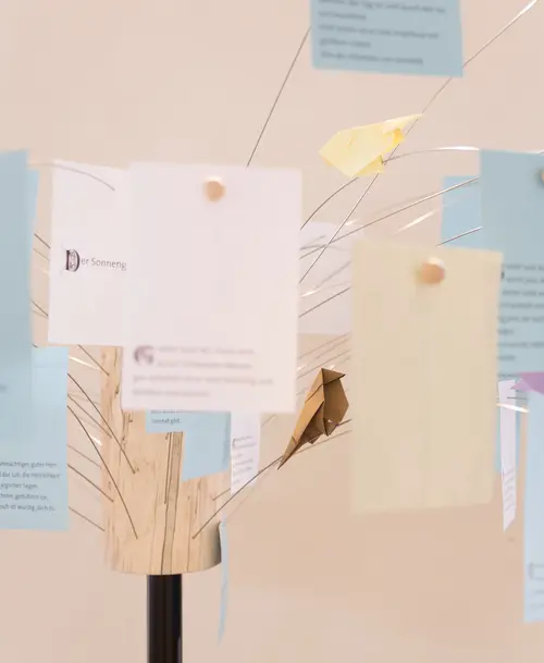 Interaktive Installation mit einem stilisierten Baum aus Holz und Draht, an dem handgeschriebene Notizen und gefaltete Papierobjekte befestigt sind.