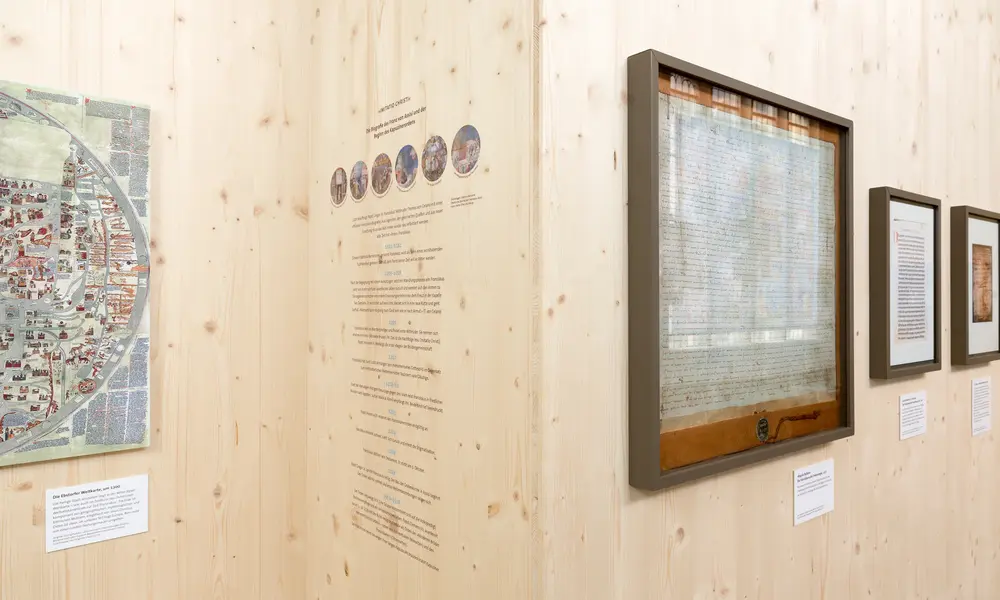 Ausstellungswand mit historischen Dokumenten, Karten und Informationstafeln, darunter eine großformatige Stadtkarte und gerahmte Schriftstücke.