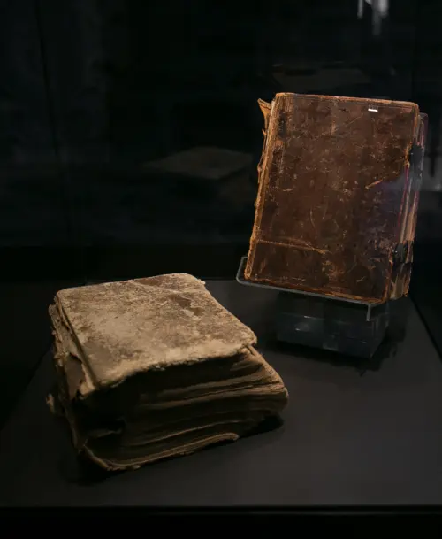 Zwei stark abgenutzte, historische Bücher mit ledergebundenen Einbänden in einer dunklen Vitrine, eines liegend und das andere aufrecht präsentiert.