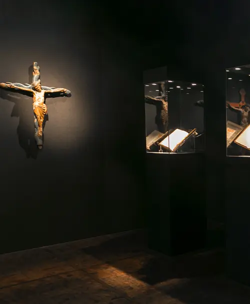 Dunkler Ausstellungsraum mit einem beleuchteten, alten Holzkruzifix an der Wand und zwei Vitrinen mit aufgeschlagenen historischen Büchern.