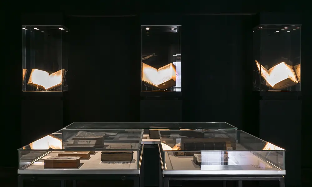 Dunkler Ausstellungsraum mit beleuchteten Vitrinen, die aufgeschlagene historische Bücher präsentieren, und einer zentralen Glasvitrine mit weiteren alten Schriftstücken.