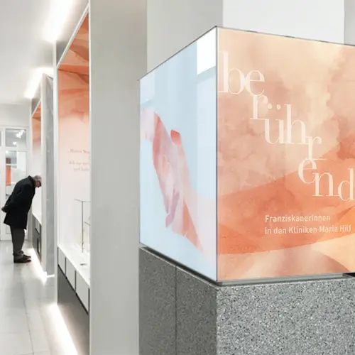 Moderne Ausstellung mit einer beleuchteten, orange-weiß gestalteten Infotafel mit dem Wort "berührend" und dem Text "Franziskanerinnen in den Kliniken Maria Hilf", während eine Person eine Vitrine betrachtet.