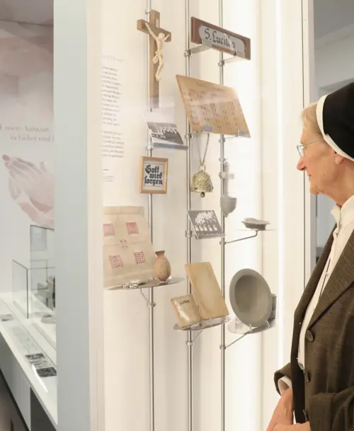 Eine Ordensschwester betrachtet eine beleuchtete Vitrine mit historischen und religiösen Objekten, darunter ein Kreuz, Bilder, Schriften und Alltagsgegenstände.