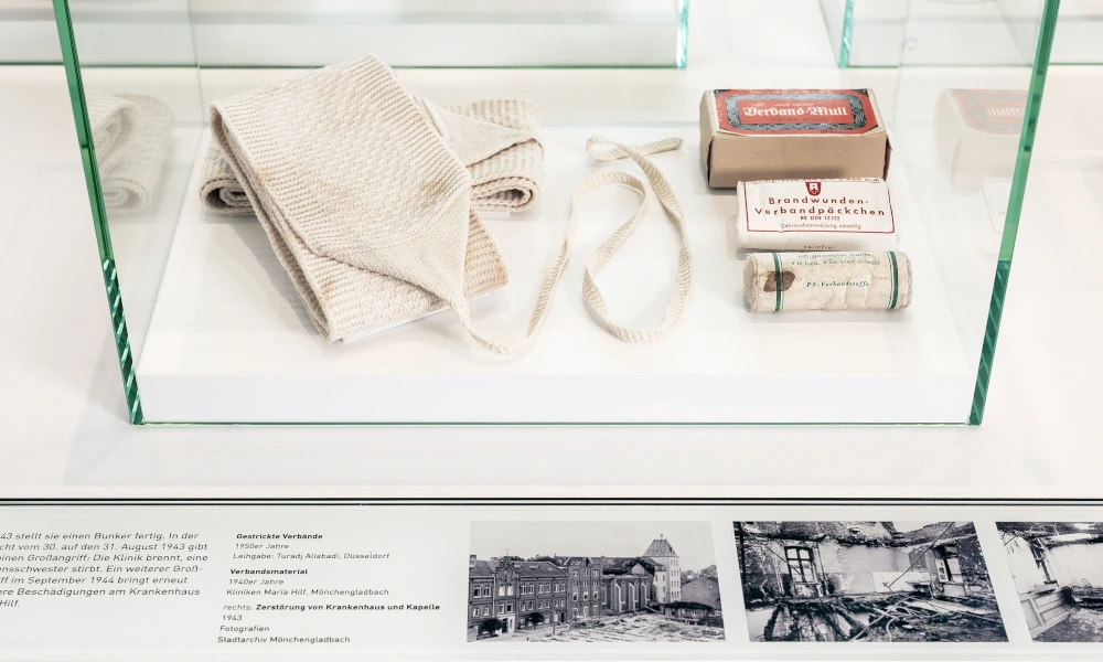 Glasvitrine mit historischen medizinischen Hilfsmitteln, darunter Verbände, Mullbinden und eine Schachtel mit Verbandwatte, begleitet von einer Informationstafel mit alten Schwarz-Weiß-Fotos von Kriegsschäden.