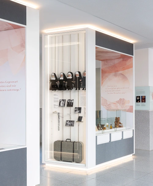 Beleuchtete Ausstellungsvitrine mit Nonnenfiguren, historischen Fotos, einem Rosenkranz, einem Koffer und weiteren Erinnerungsstücken, umgeben von modern gestalteten Informationstafeln.