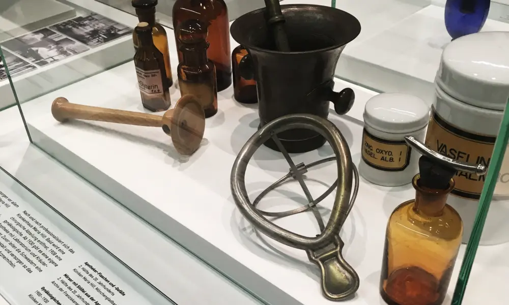 Glasvitrine mit historischen Apotheken- und Medizinutensilien, darunter Glasfläschchen, ein Mörser mit Stößel, eine Salbenschale und ein metallisches medizinisches Instrument.