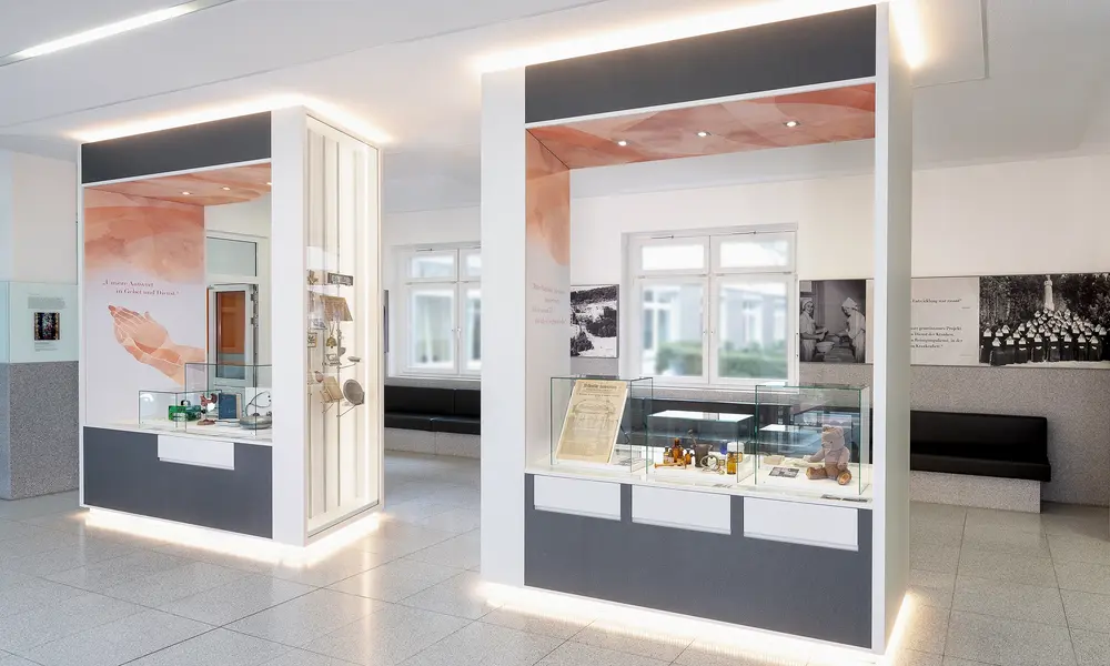 Moderner Ausstellungsraum mit beleuchteten Vitrinen, historischen Exponaten und Informationstafeln, ergänzt durch große Fenster und eine helle, offene Gestaltung.