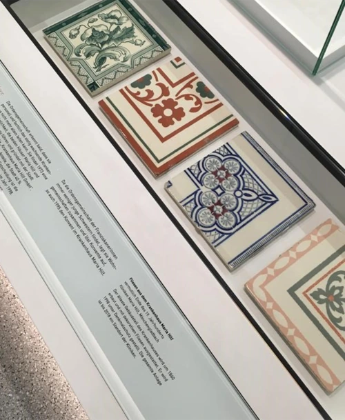 Glasvitrine mit vier historischen Fliesen in verschiedenen Farben und Mustern, begleitet von einer Informationstafel mit Erläuterungen zur Herkunft und Bedeutung.