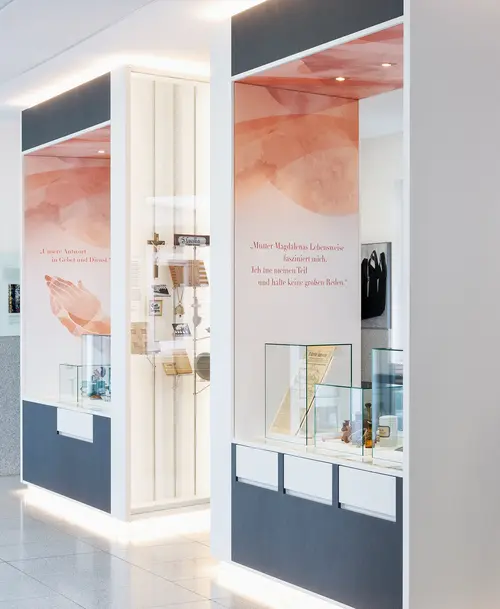 Moderner Ausstellungsraum mit beleuchteten Vitrinen, historischen Exponaten und Informationstafeln mit Zitaten, gestaltet in hellen Farben und klaren Linien.