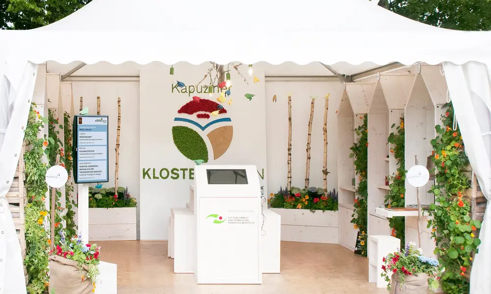 Weißes Ausstellungszelt mit dem Schriftzug „Kapuziner KLOSTER“ auf der Rückwand, begrünten Elementen, Infotafeln und einem zentralen Touchscreen-Display.