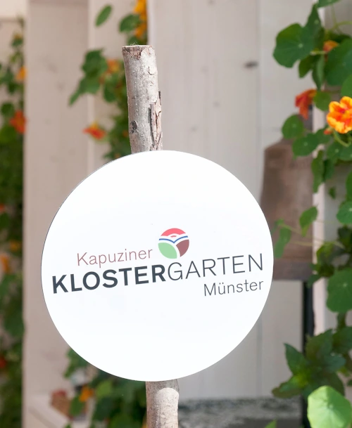 Rundes Schild mit der Aufschrift „Kapuziner KLOSTERGARTEN Münster“, befestigt an einem Holzstab, vor einer begrünten Kulisse mit orangefarbenen Blüten.