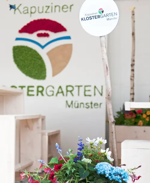 Ausstellungsbereich des „Kapuziner KLOSTERGARTEN Münster“ mit einem runden Schild auf einem Holzstab, bunten Blumen im Vordergrund und einer begrünten Logo-Wand im Hintergrund.